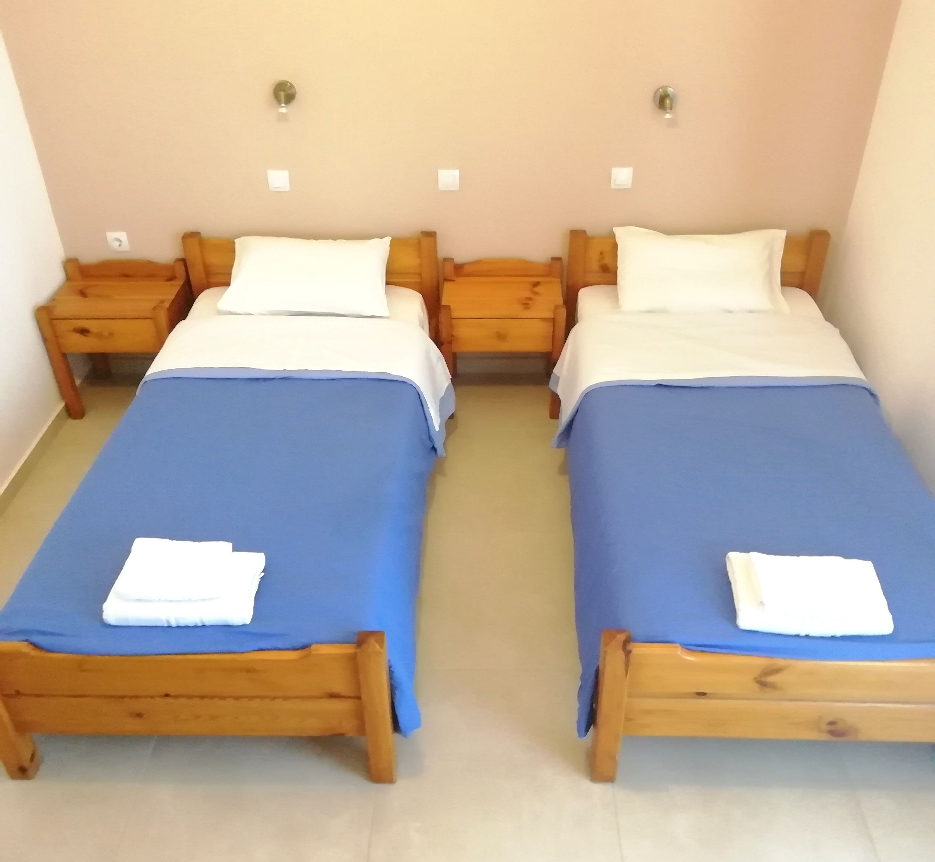 A room with two wooden beds, each with a blue bedspread and white pillows, and a small wooden nightstand beside each bed.