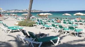 A beach with rows of green lounge chairs and umbrellas, a palm tree, and a clear view of the sea and distant buildings.