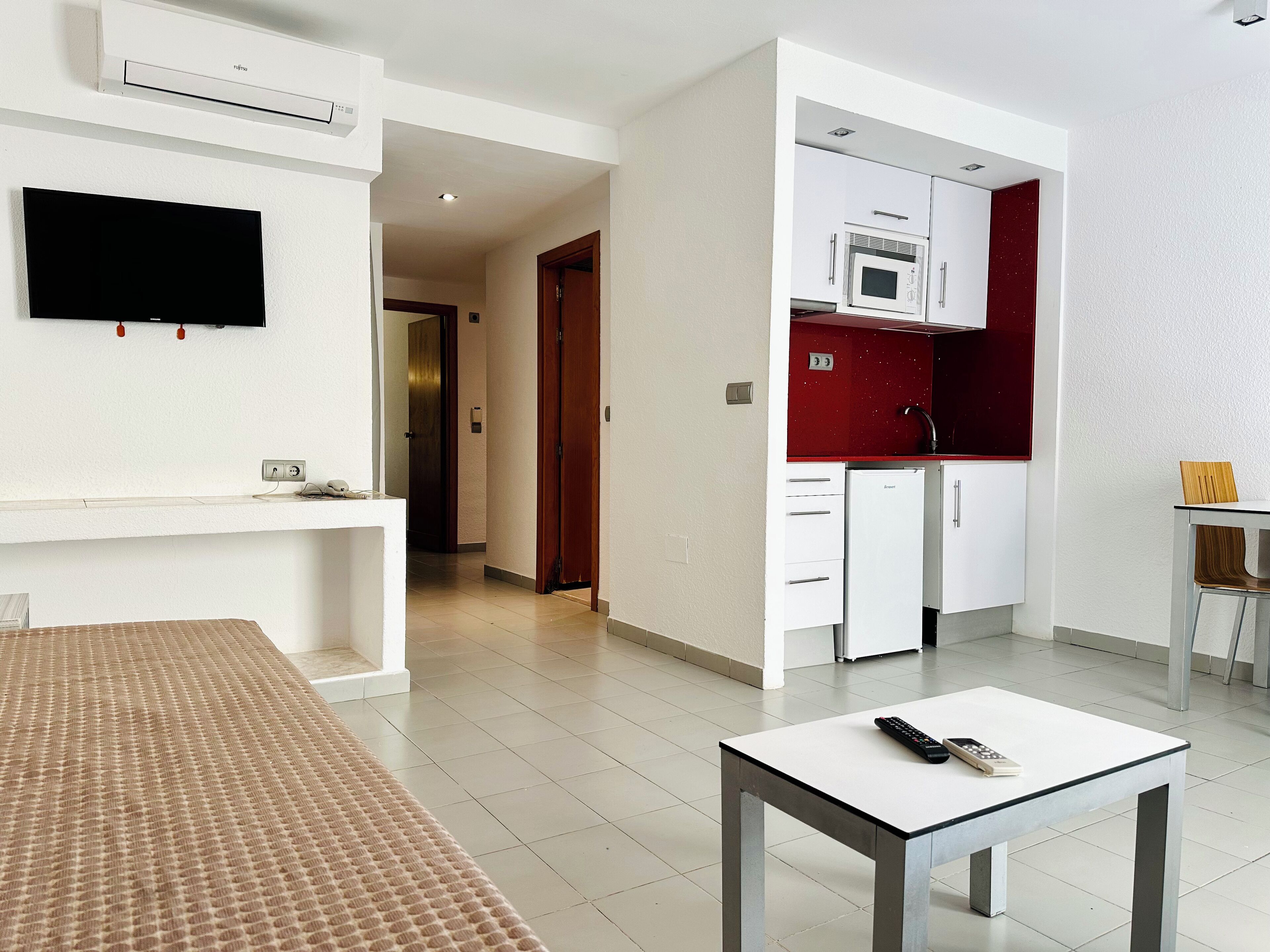 A hotel room with a bed, a TV mounted on the wall, a kitchenette with a microwave, and a small table with a remote control.