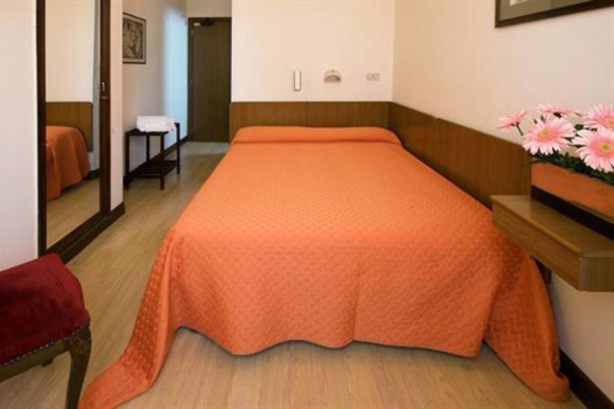 Habitación individual con una cama de una sola cama, cabecera de madera, mesita de noche pequeña, una silla roja y un florero sobre la mesita.
