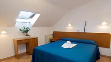 Une chambre avec un puits de lumière, un lit recouvert d’une courtepointe bleue, un bureau en bois, une plante en pot et deux lampes fixées au mur.