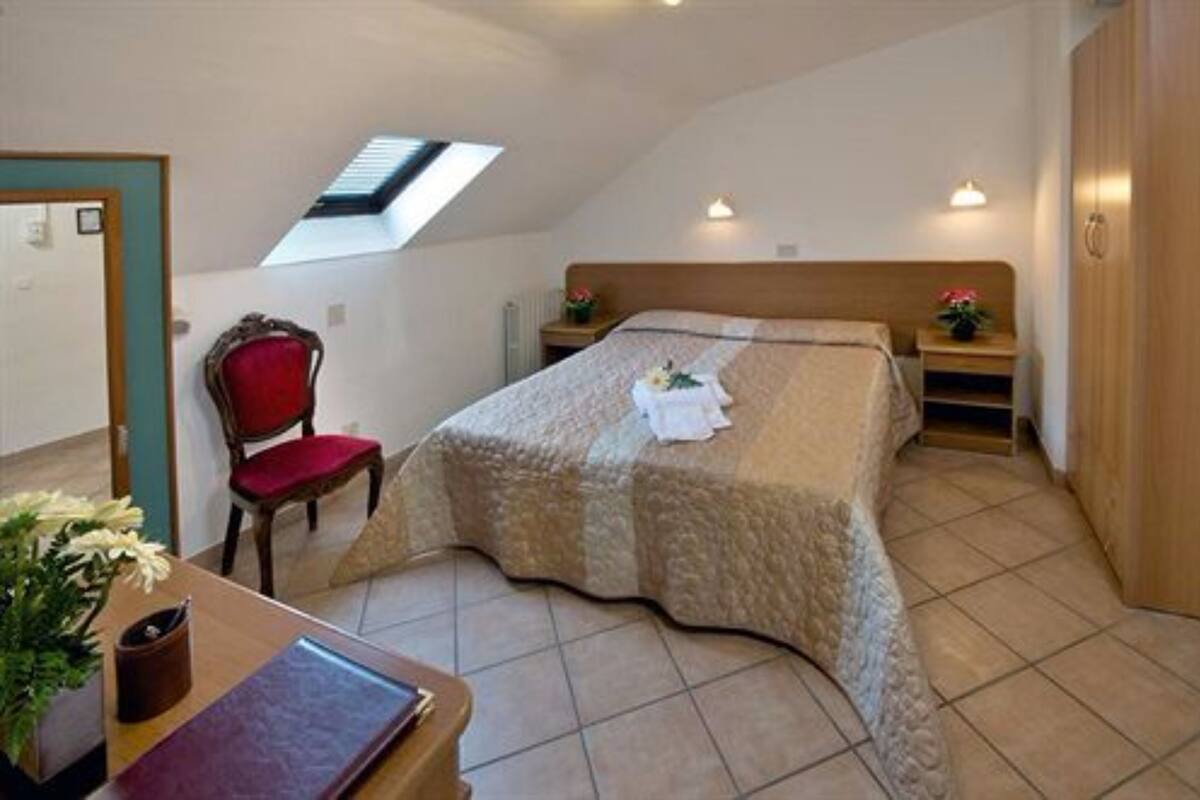 Un dormitorio con cama, una silla roja, una mesita con un florero, un tragaluz y un armario de madera.