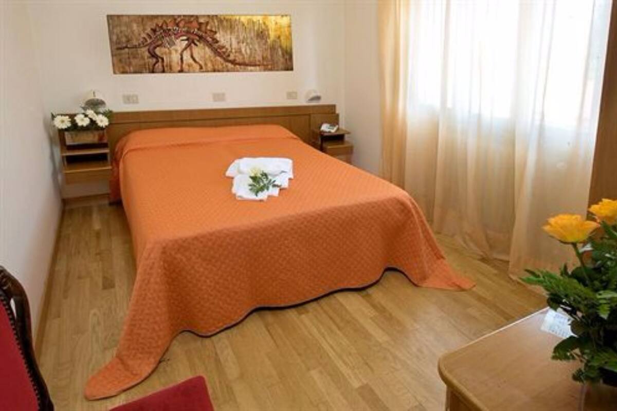 Una cama bien hecha con una colcha naranja, una mesita de noche con lámpara, un cuadro colgado en la pared y una ventana con cortinas.