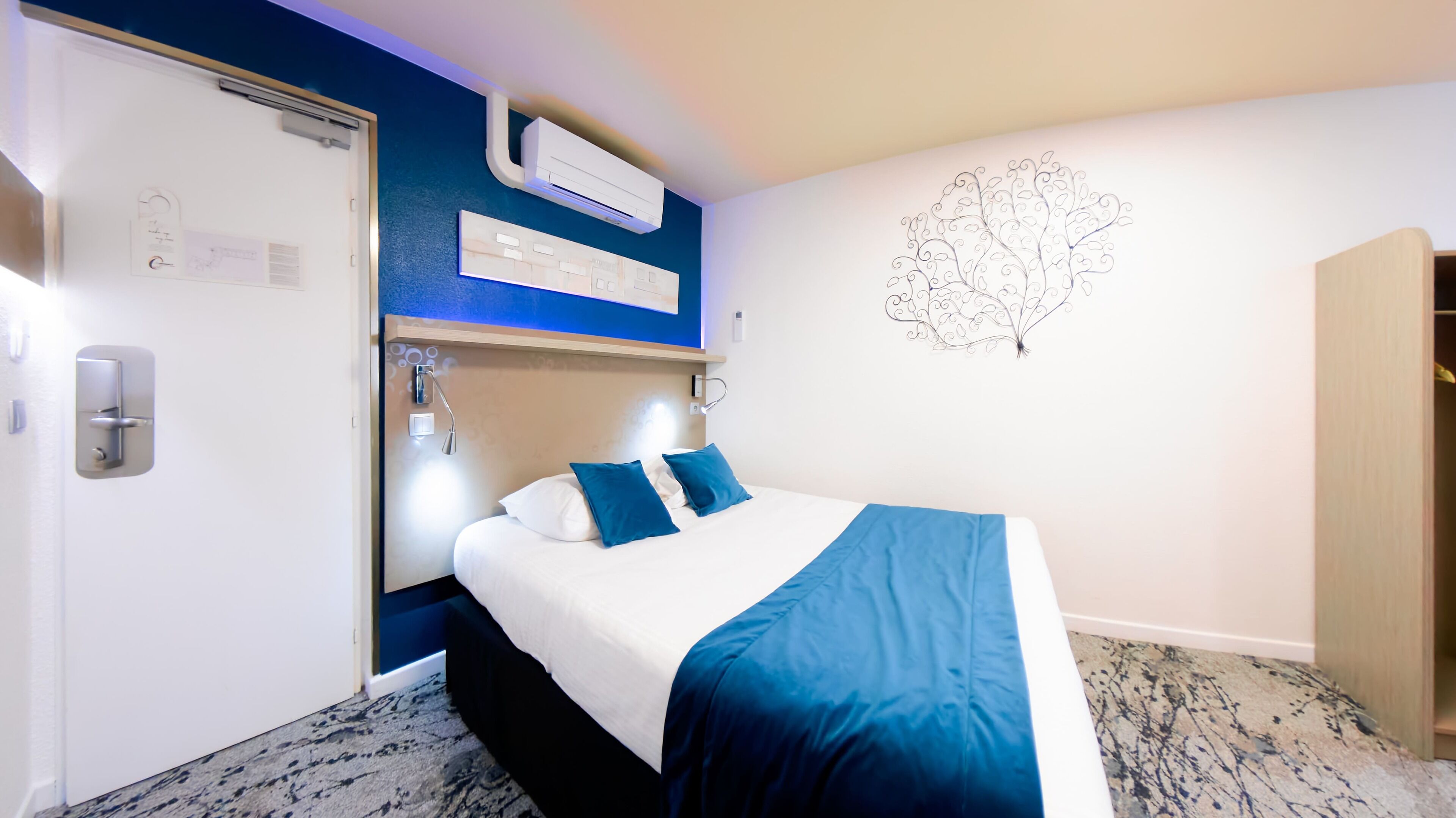 A hotel room with a bed, a nightstand, a wall-mounted air conditioner, a door with a handle, a light switch, and a wall decoration.
