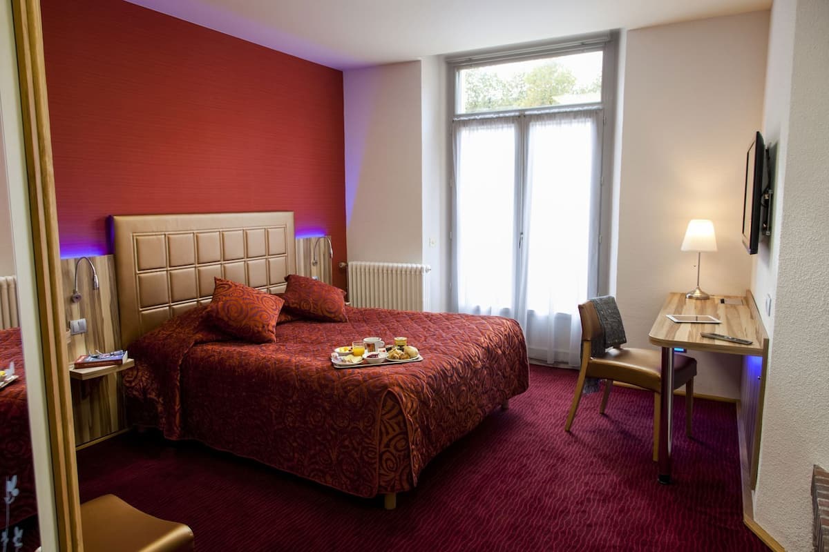 A hotel room with a large bed, a bedside table with a lamp, a chair, and a window with curtains.