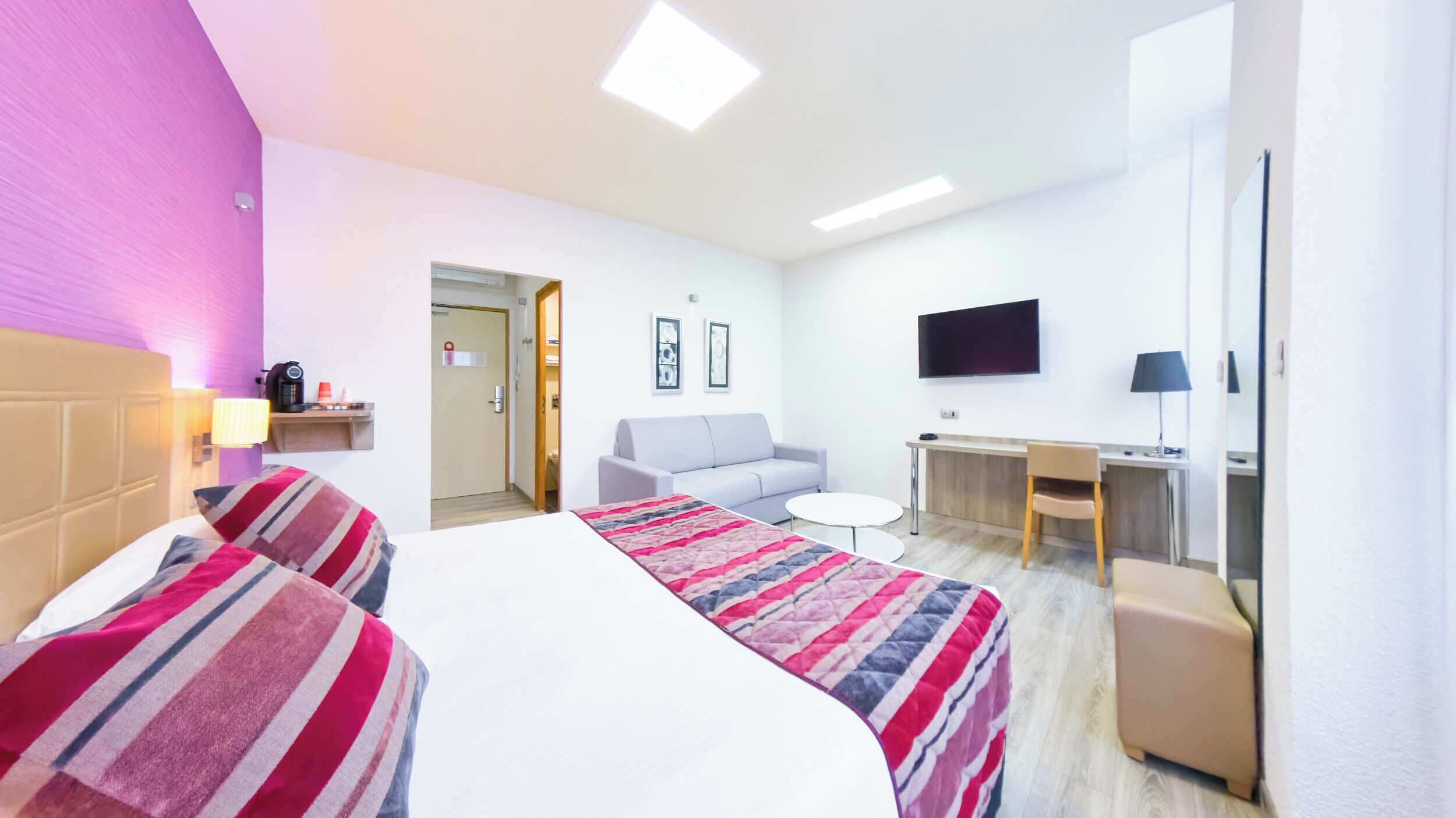 A hotel room with a bed, two bedside tables, a sofa, a desk, a TV, and a chair.