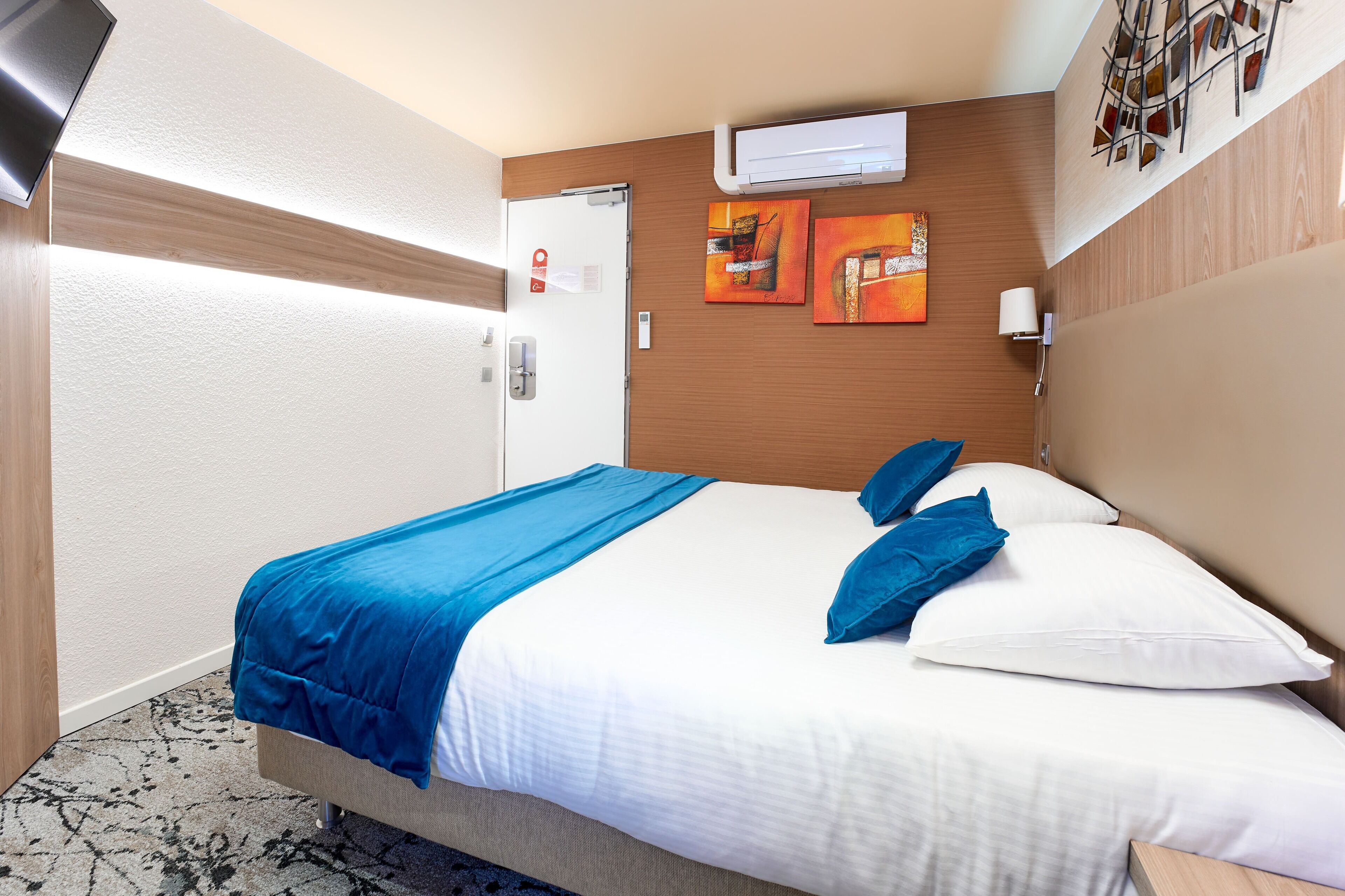 Une chambre d’hôtel avec un lit, une literie bleue et blanche, une tête de lit en bois, un climatiseur fixé au mur et deux tableaux.