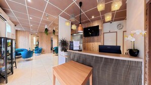 Ein Hotel-Empfangsbereich mit Holzbüro, blauen Sesseln, einer Holzwand, einer Uhr und einem an der Wand montierten Fernseher.
