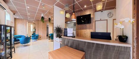 A hotel reception area with a wooden reception desk, blue chairs, a wooden wall, a clock, and a TV mounted on the wall.