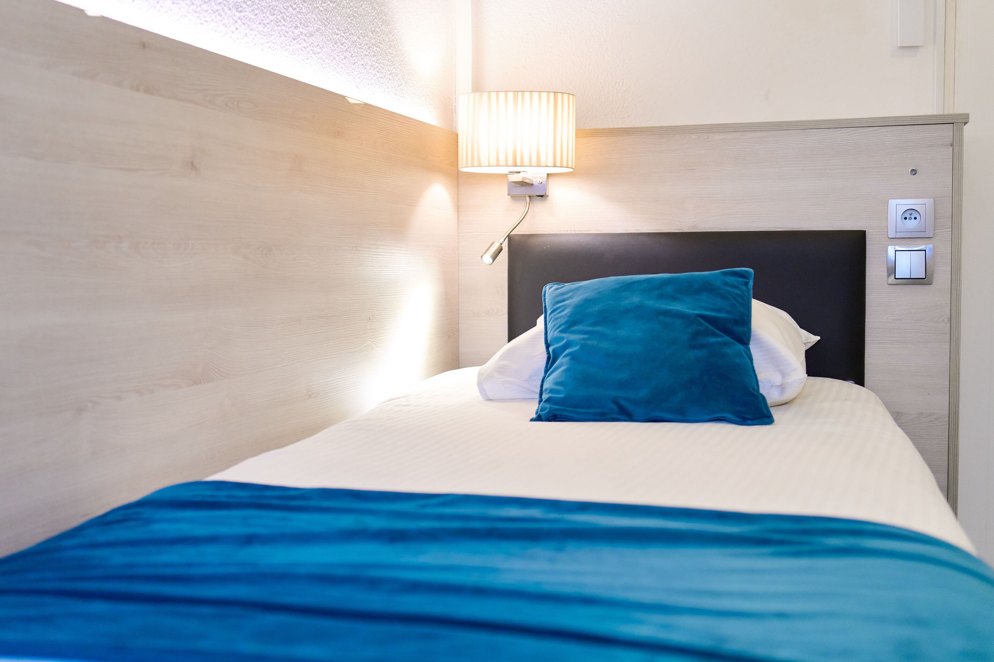 A neatly made bed with a blue pillow and a white pillow, a wall-mounted lamp, and a power outlet on the wall.