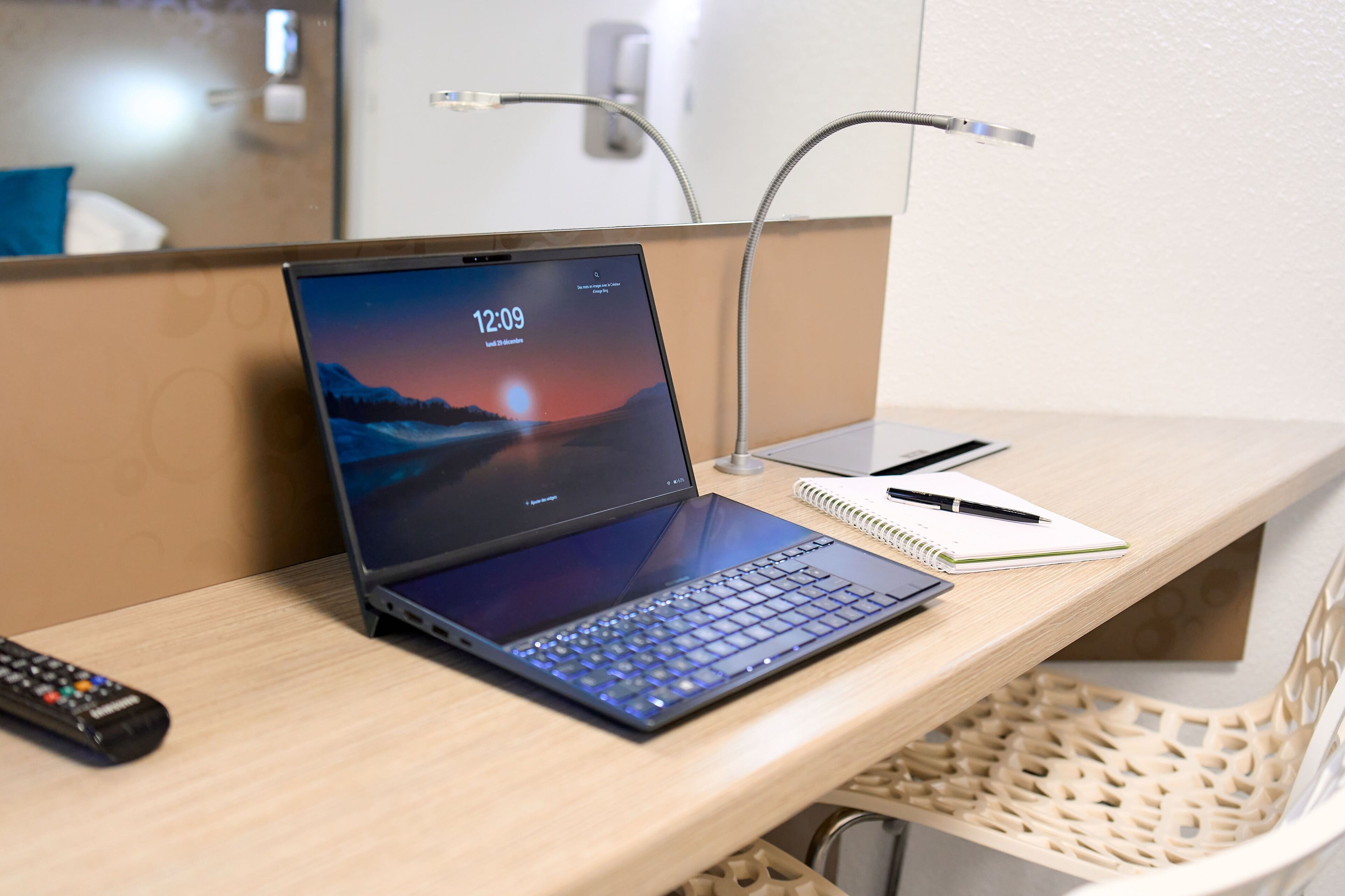 A laptop on a desk with a notebook and pen, a remote control, and a chair with a woven backrest.
