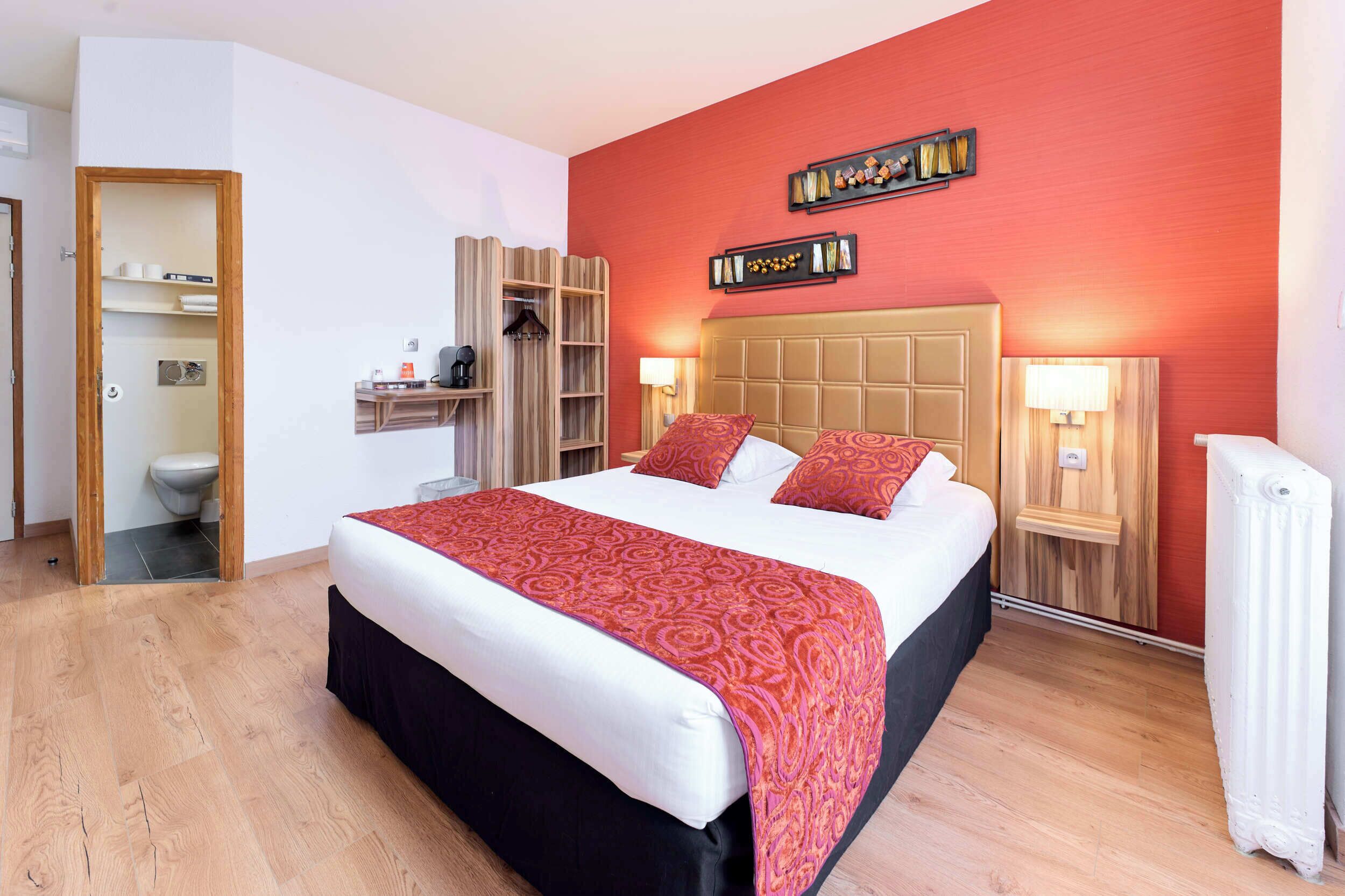 A hotel room with a large bed, wooden headboard, two bedside tables with lamps, a wardrobe, and a small shelf with toiletries.