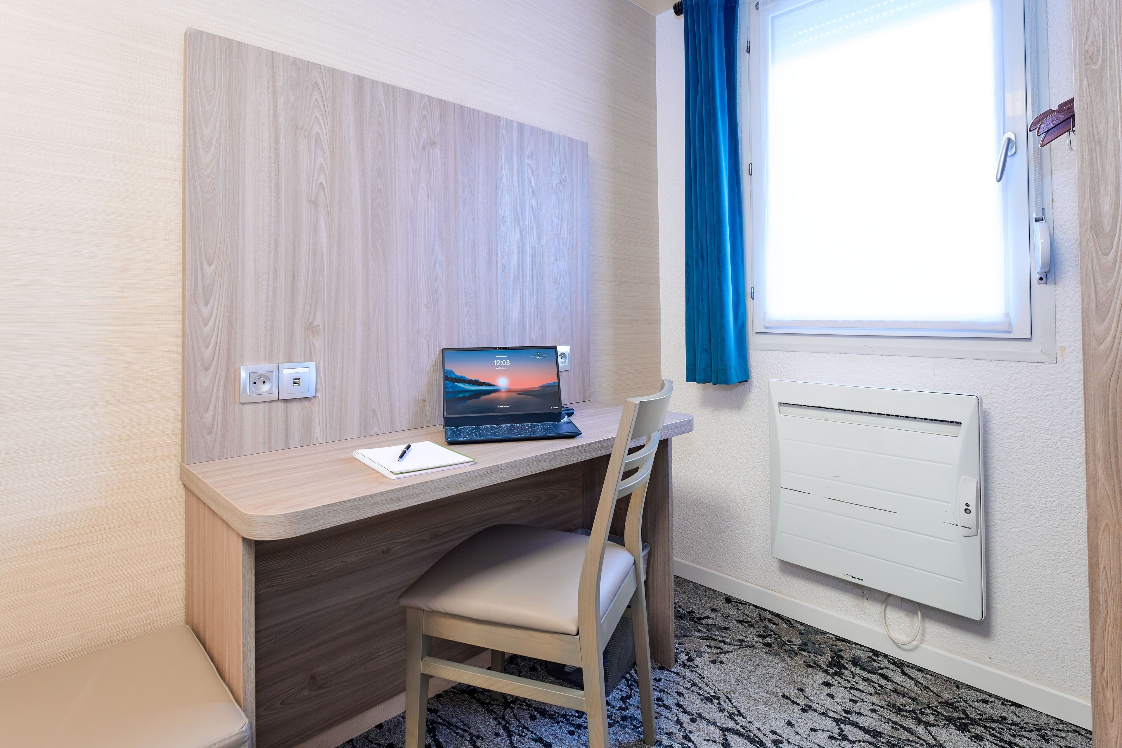 A modern room with a wooden desk, a laptop, a chair, a window with blue curtains, and a wall-mounted air conditioner.