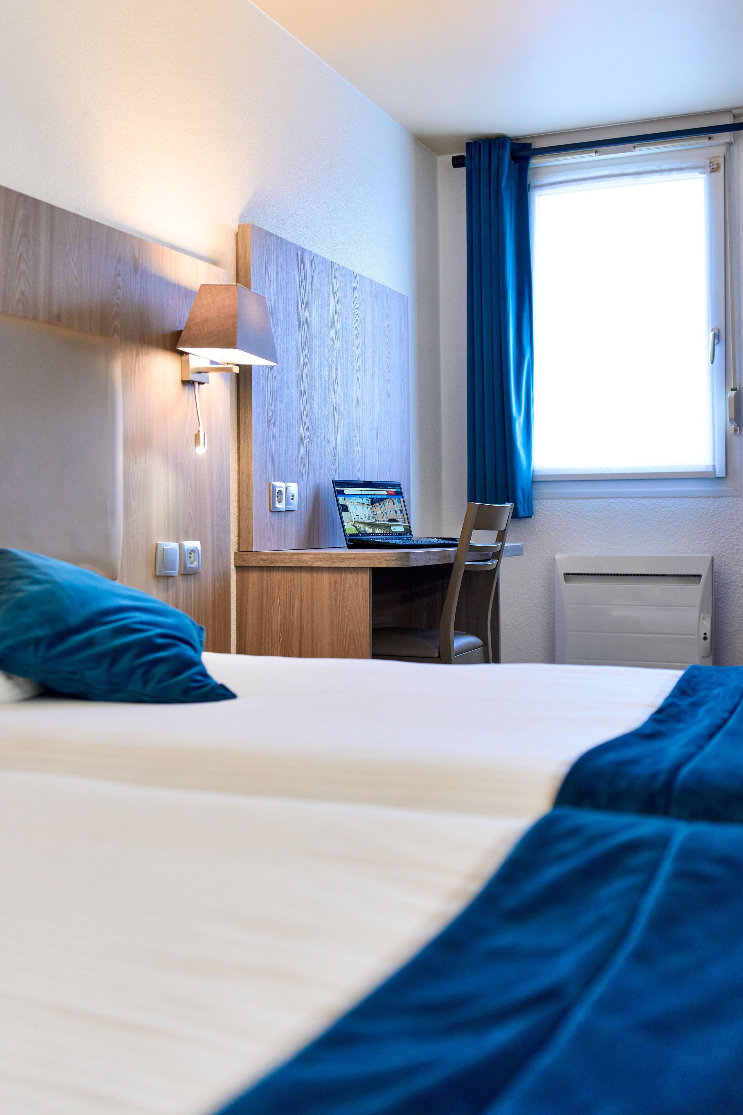 A hotel room with a bed, a desk with a laptop, a chair, a lamp, and a window with curtains.