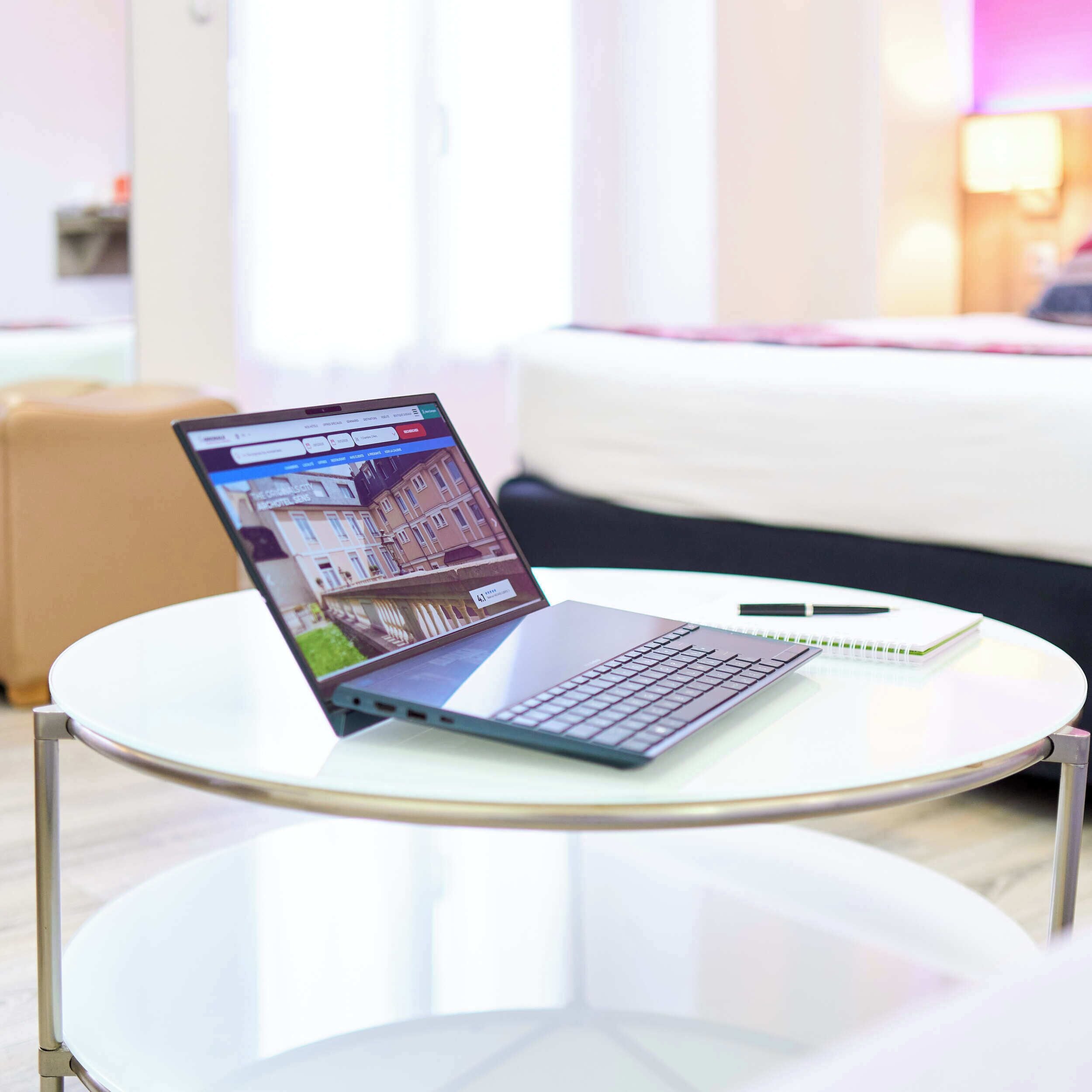 Een laptop op een glazen tafel, met een notitieboekje en pen ernaast.