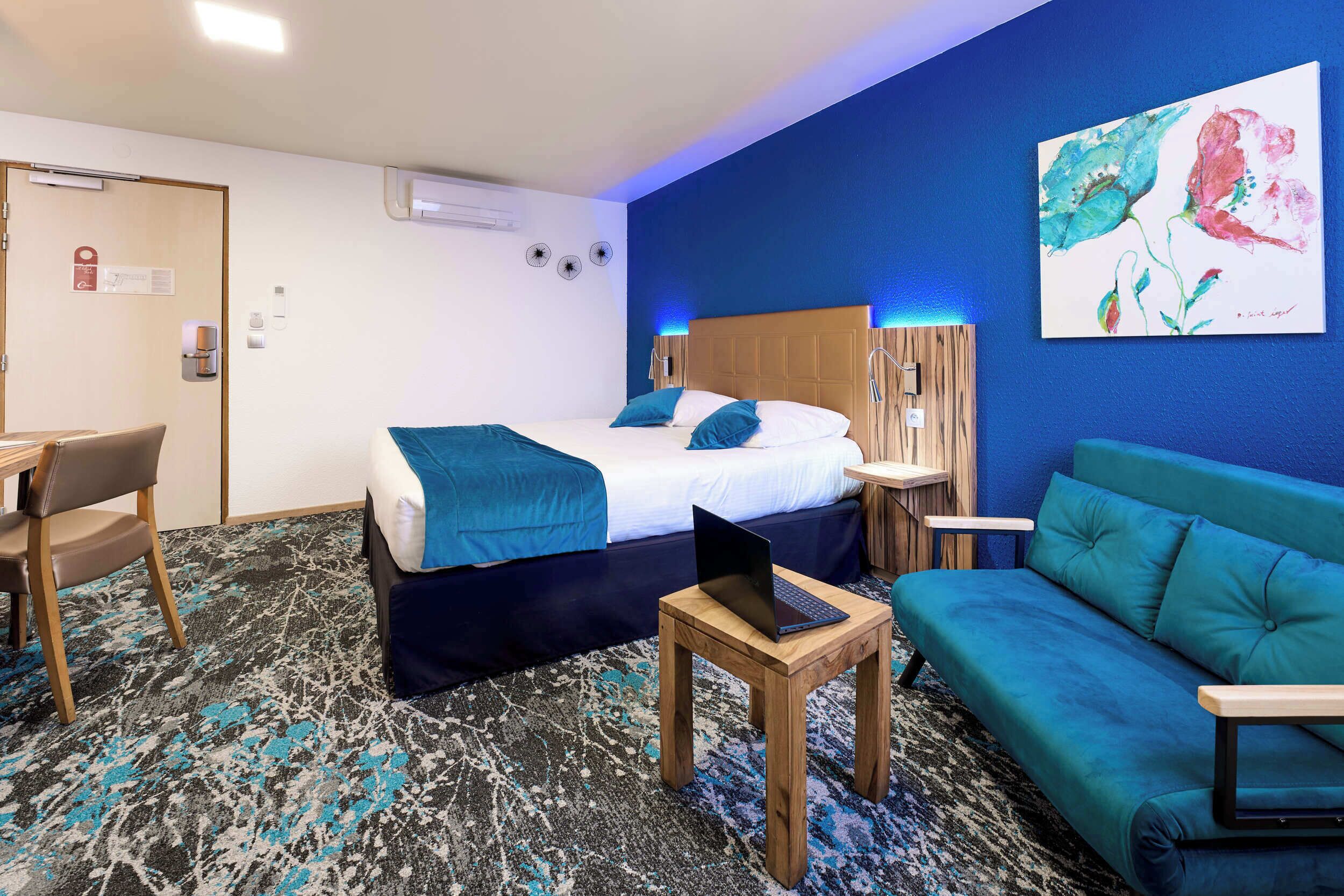 A hotel room with a bed, a desk, a laptop, a sofa, and a painting on the wall.