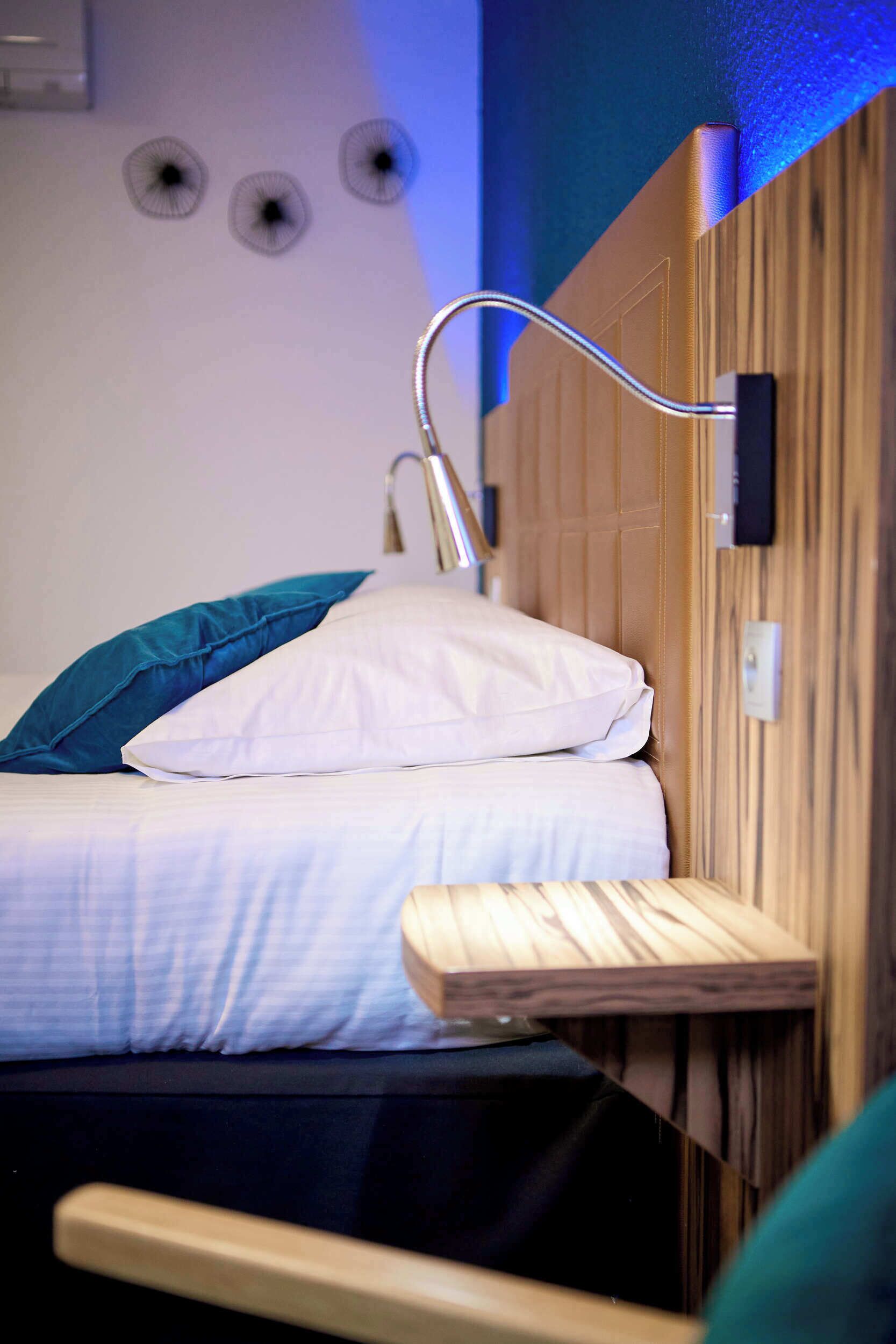 Una cama individual con cabecera de madera, un almohadón blanco y un almohadón color turquesa. Se observan tres rejillas circulares fijadas a la pared sobre la cama.