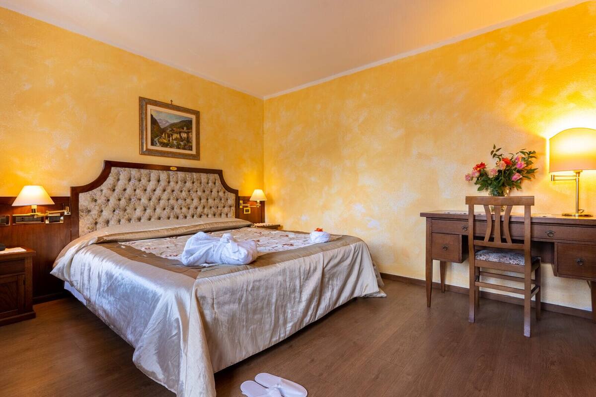 A hotel room with a large bed, two bedside lamps, a wooden desk, a chair, and a painting on the wall.