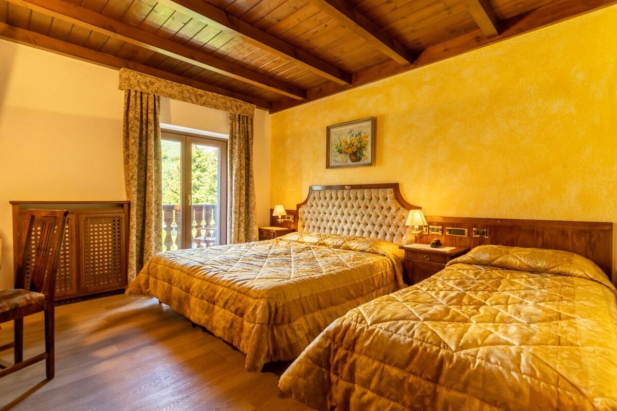 A hotel room with two beds, wooden ceiling beams, a window with curtains, a nightstand with a lamp, and a framed picture on the wall.