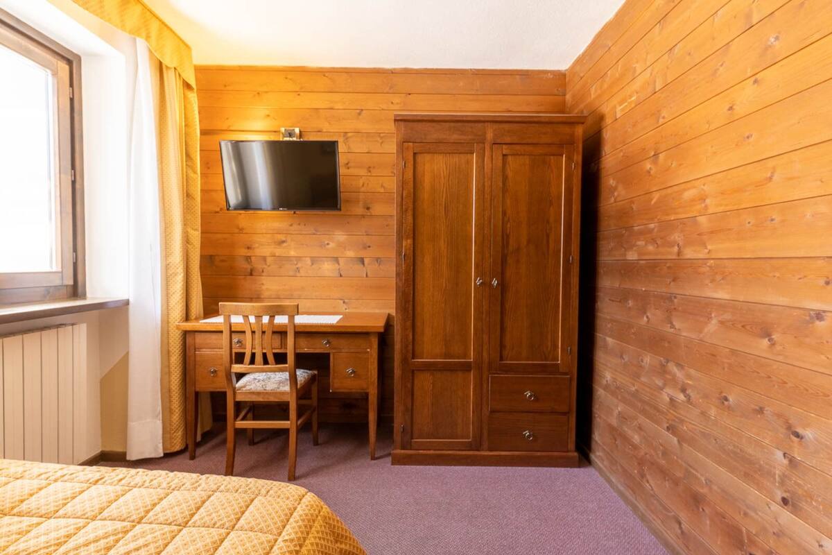 A room with wooden panel walls, a bed with a yellow quilt, a wooden desk with a chair, a television mounted on the wall, and a wardrobe.