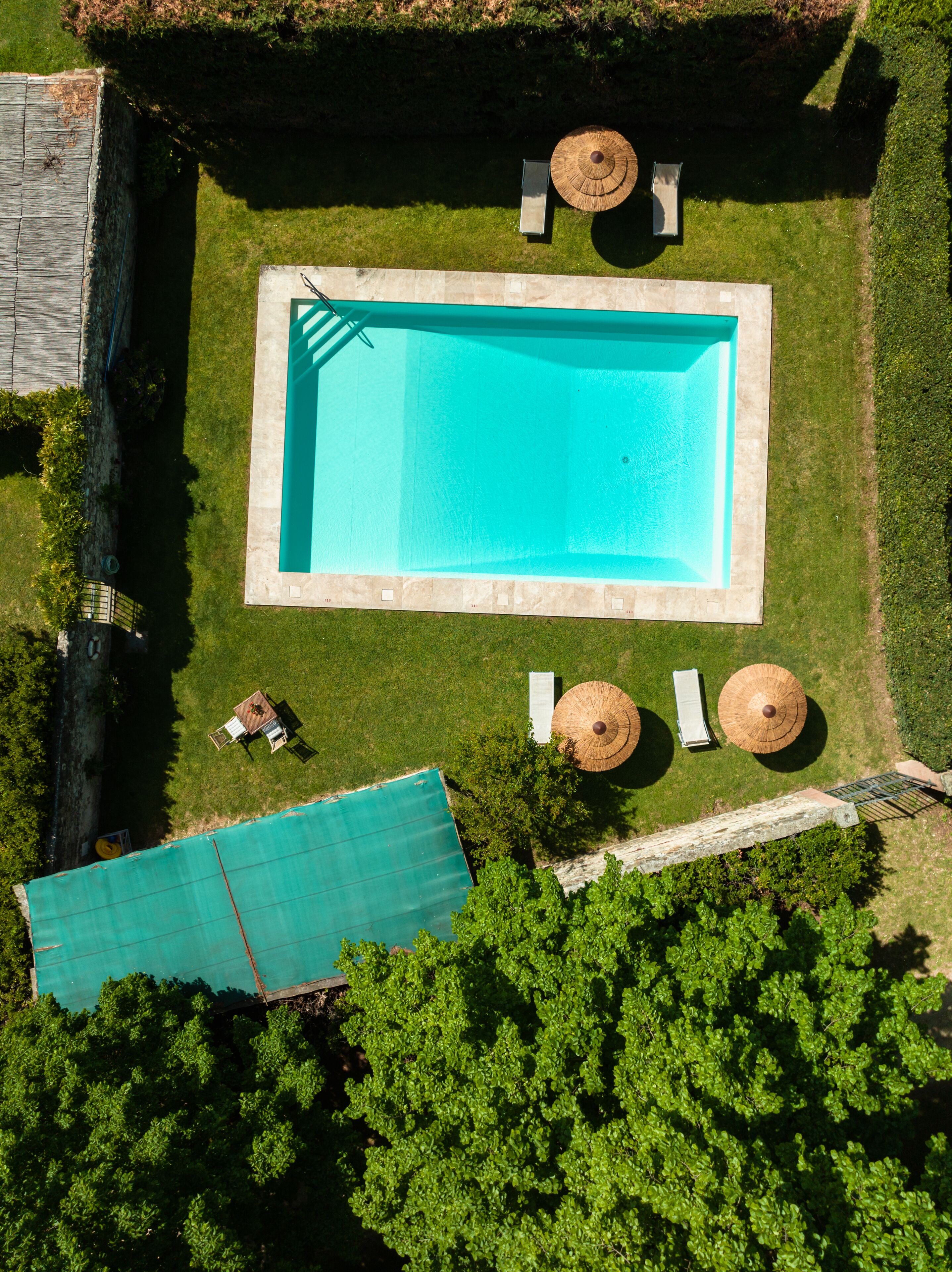 Vista aérea de una piscina rectangular rodeada por una terraza de piedra, con dos sillas de descanso y dos sombrillas de sol.