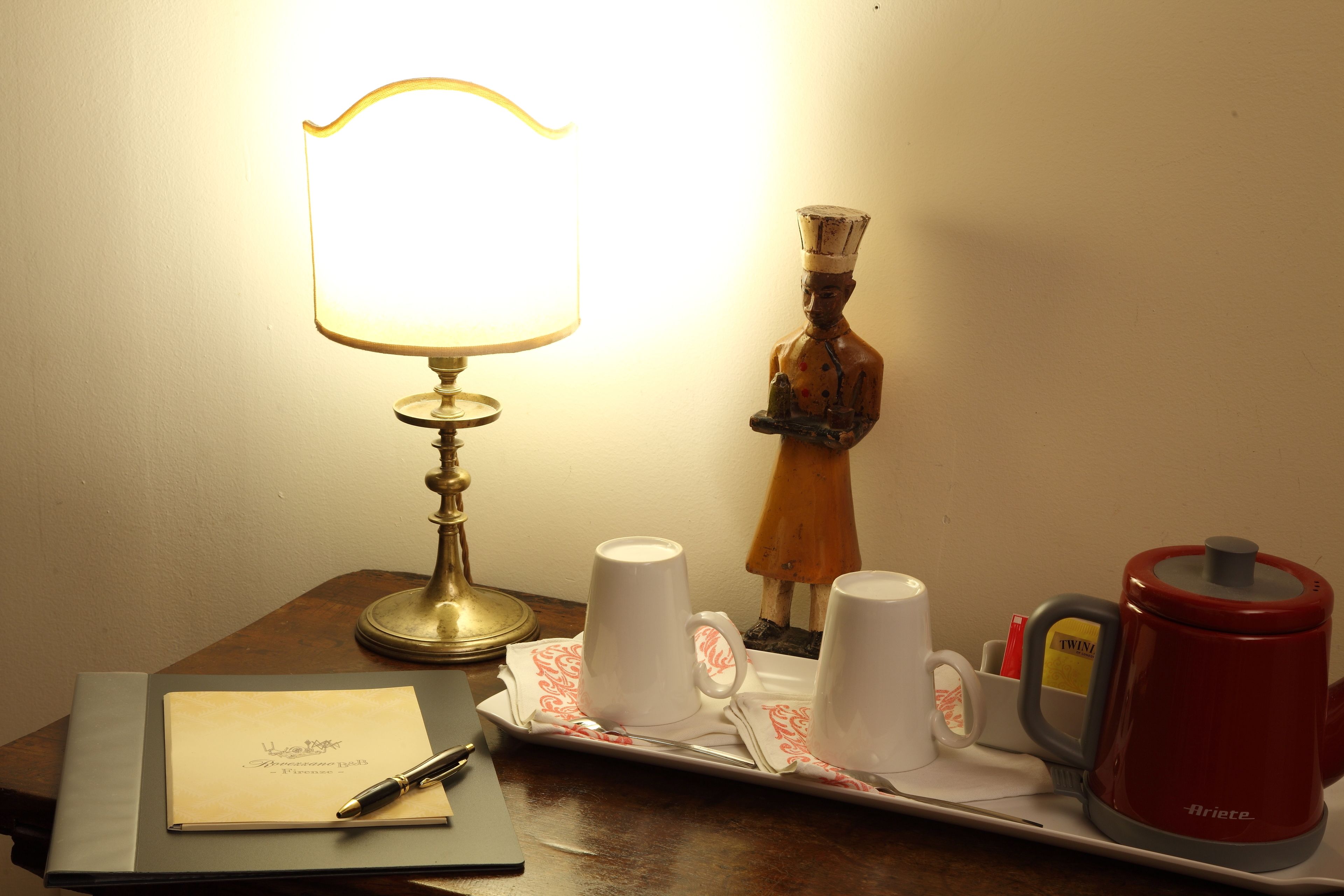 A table with a lamp, a tray with cups, a figurine, and a red kettle.
