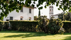 A white building with a tower, surrounded by a well-maintained hedge and palm trees.