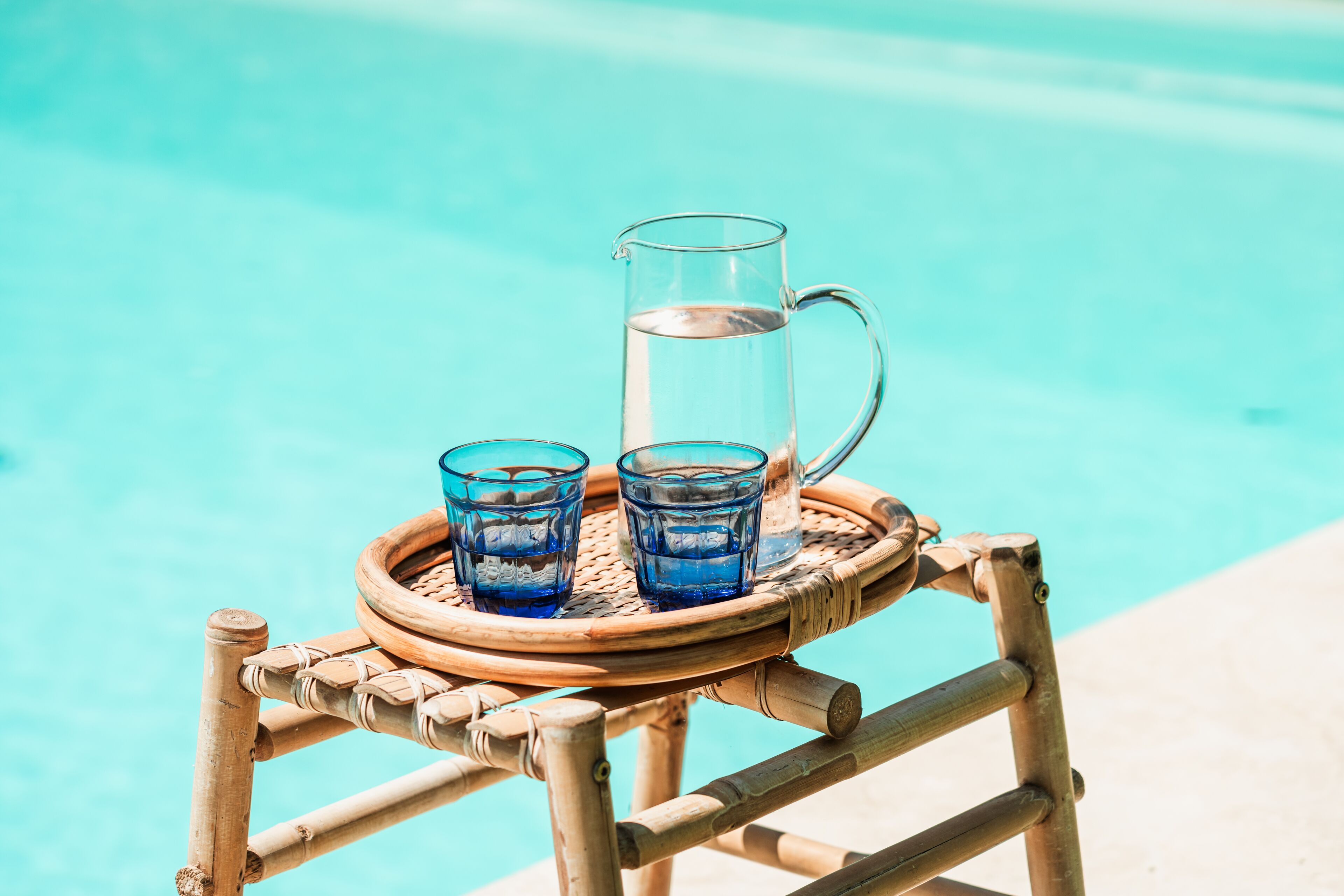 A bamboo table with a glass pitcher and two glasses of water by a pool.