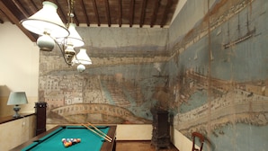 A pool table with balls, a lamp, and a chair in a room with a mural of a cityscape and ships on the wall.