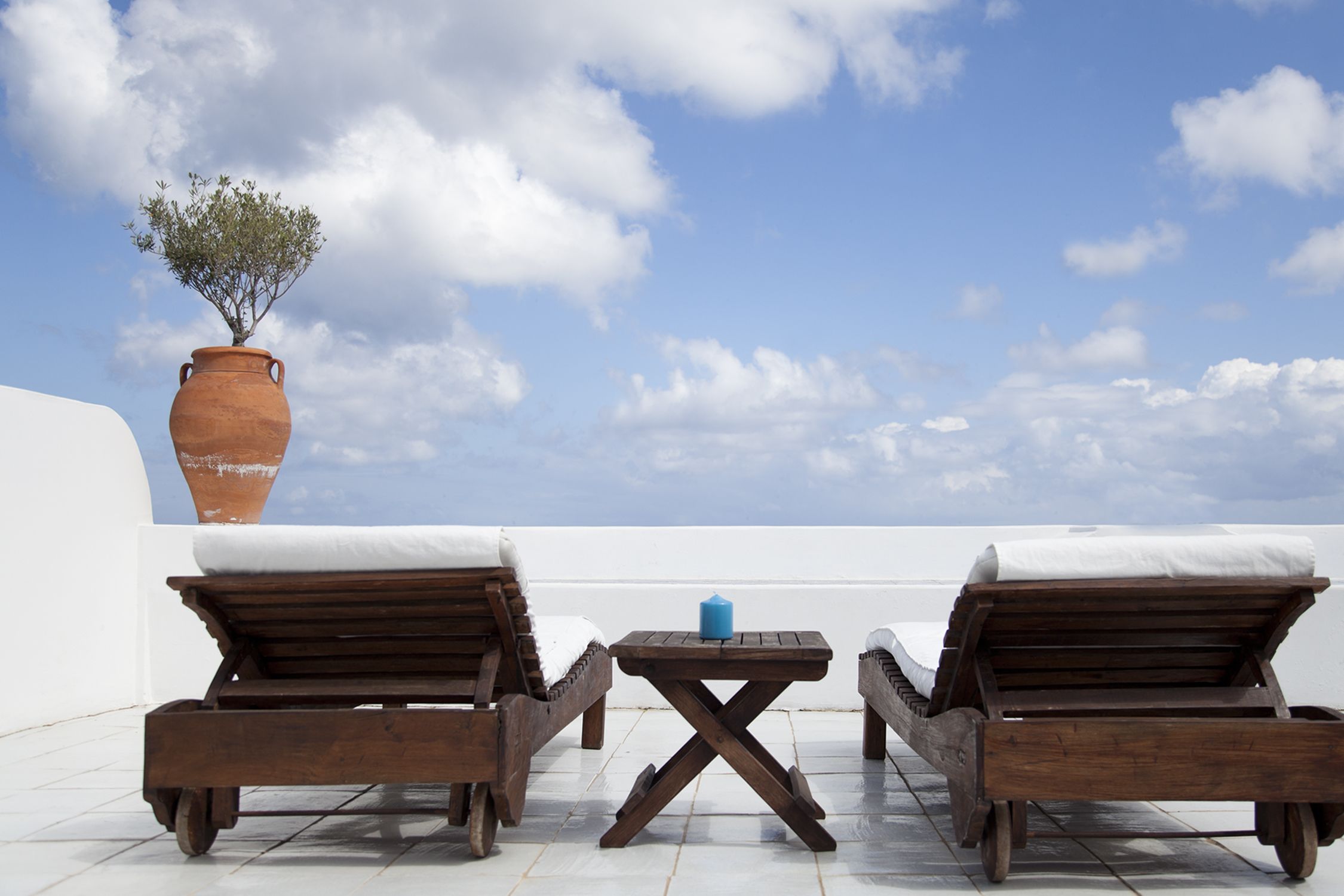 Two wooden lounge chairs with cushions, a small table with a candle, and a large terracotta pot with a plant.