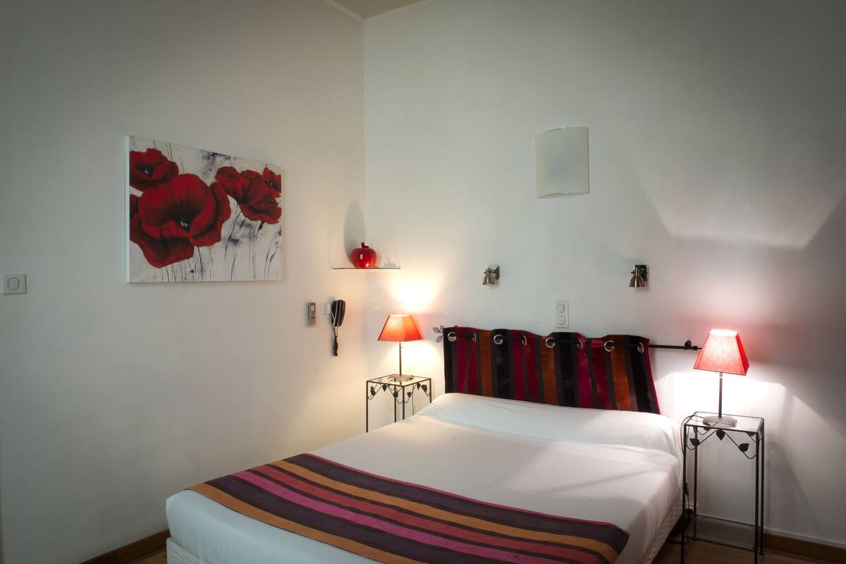 A hotel room with a bed, two bedside lamps, a red flower painting, a small shelf, and a wall-mounted light switch.