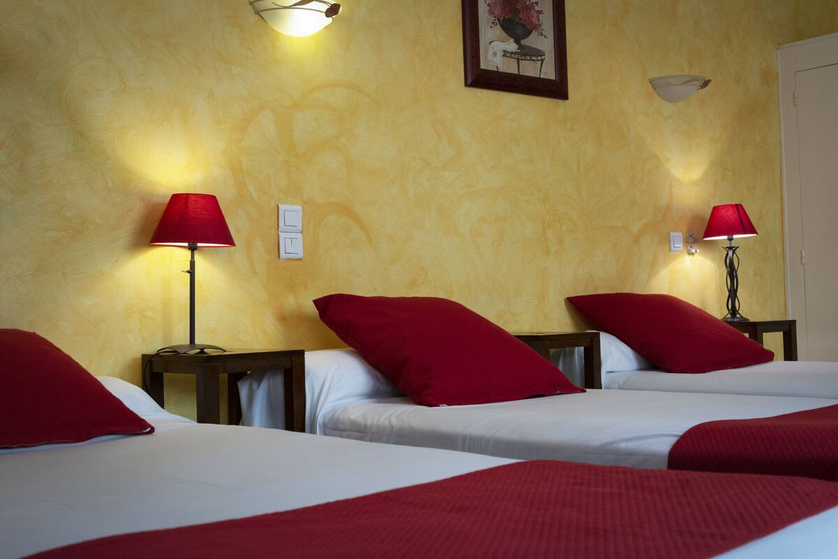 A hotel room with three beds, each with red pillows and white bedding. There are two wall-mounted lamps with red shades.