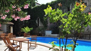 Zona junto a la piscina con mobiliario de madera y un edificio de piedra al fondo.