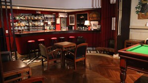 Zona de bar con mesa de billar, sillas de madera y barra con diferentes botellas y vasos.