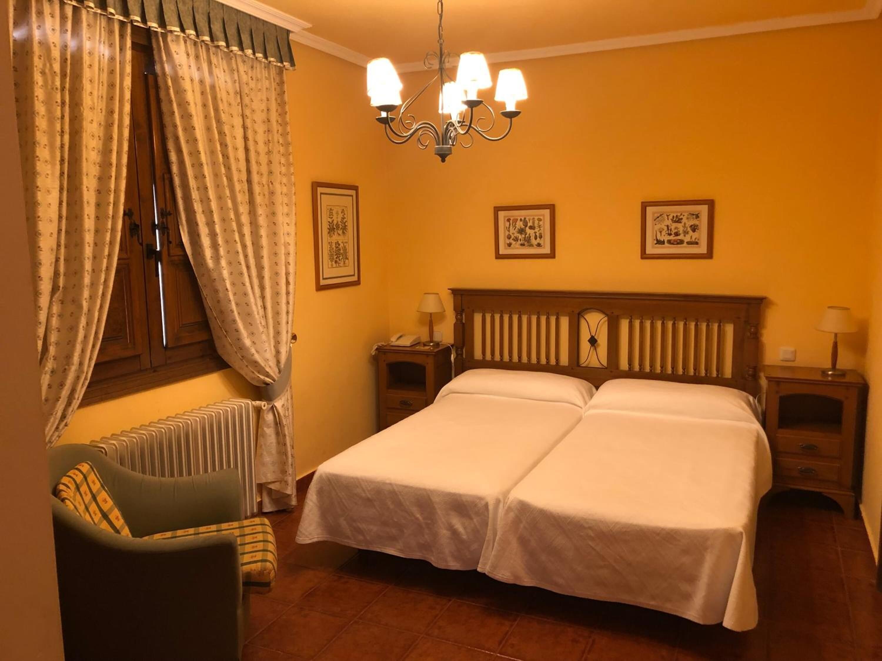 A hotel room with two beds, a chair, a radiator, and a chandelier.