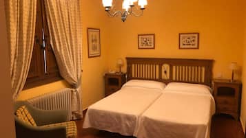 A hotel room with two beds, a chair, a radiator, and a chandelier.