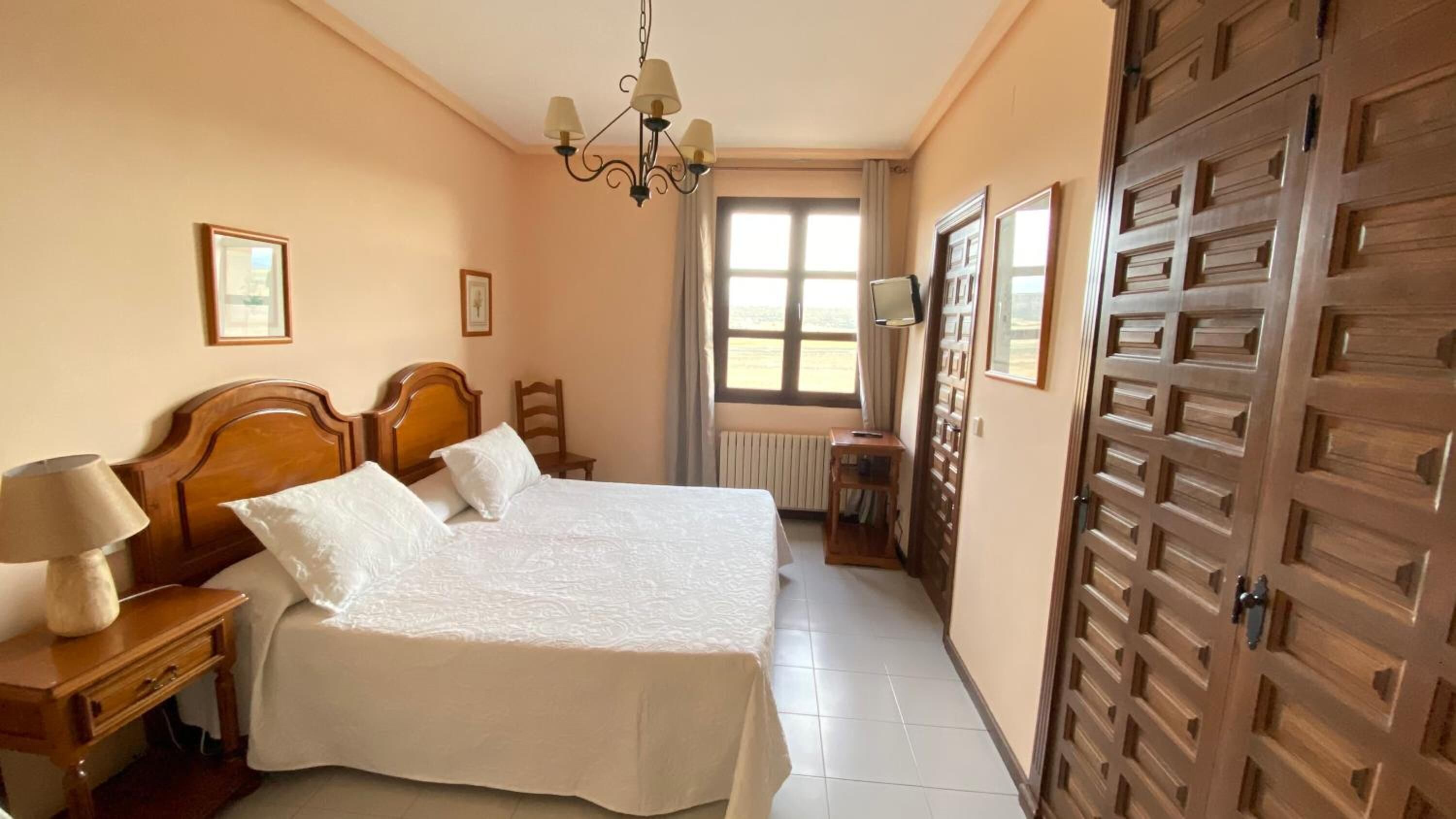 A bedroom with two beds, a chandelier, a window with curtains, and a wooden wardrobe.