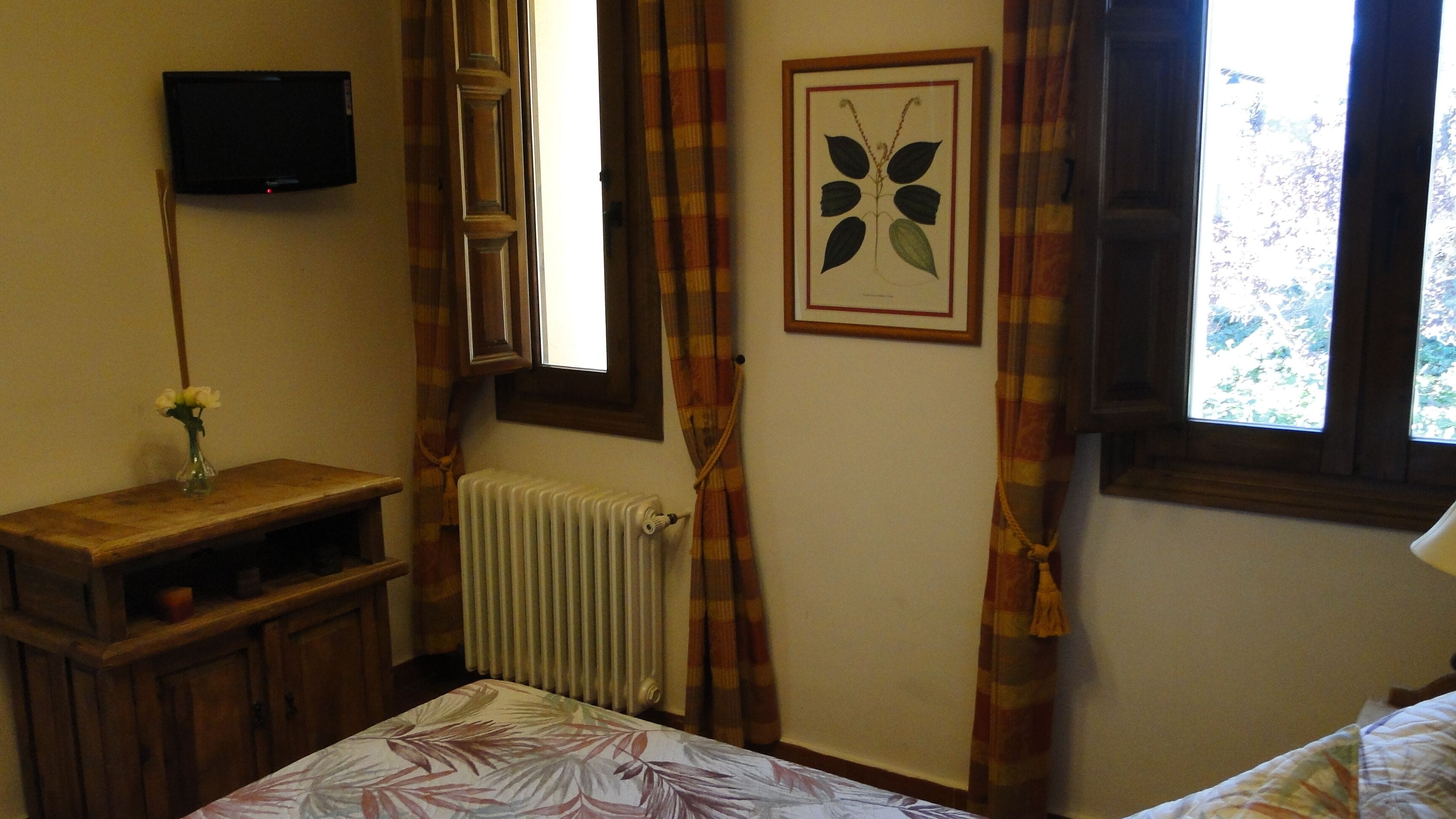 Een slaapkamer met een bed, een nachtkastje met een bloevas, een radiator, een ingelijste botanische print aan de muur, een aan de muur bevestigde televisie, en een raam met gordijnen.