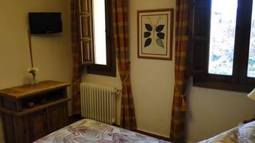 Een slaapkamer met een bed, een nachtkastje met een bloevas, een radiator, een ingelijste botanische print aan de muur, een aan de muur bevestigde televisie, en een raam met gordijnen.
