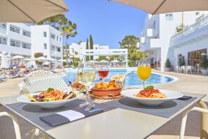 A dining table with plates of food and drinks, overlooking a pool and white buildings. - Prinsotel Alba Hotel Apartamentos (Santanyi)