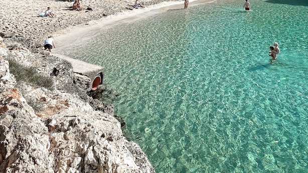 Una playa con agua turquesa cristalina, costa rocosa y edificios al fondo.