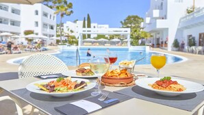 Una mesa de comedor con platos de comida, una copa de vino y un cóctel, con vistas a una zona de piscina con gente y edificios al fondo.