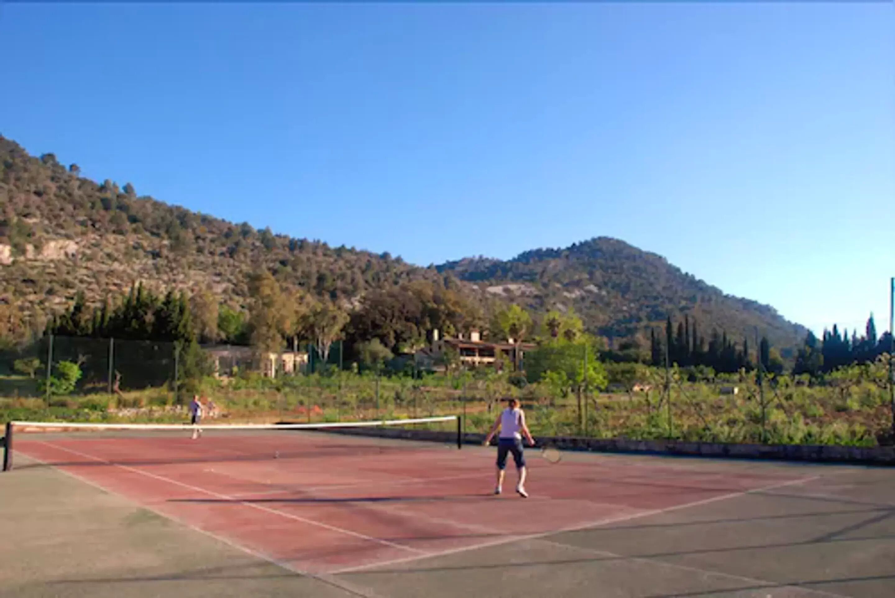 Una cancha de tenis con dos jugadores, rodeada por un paisaje montañoso y árboles.