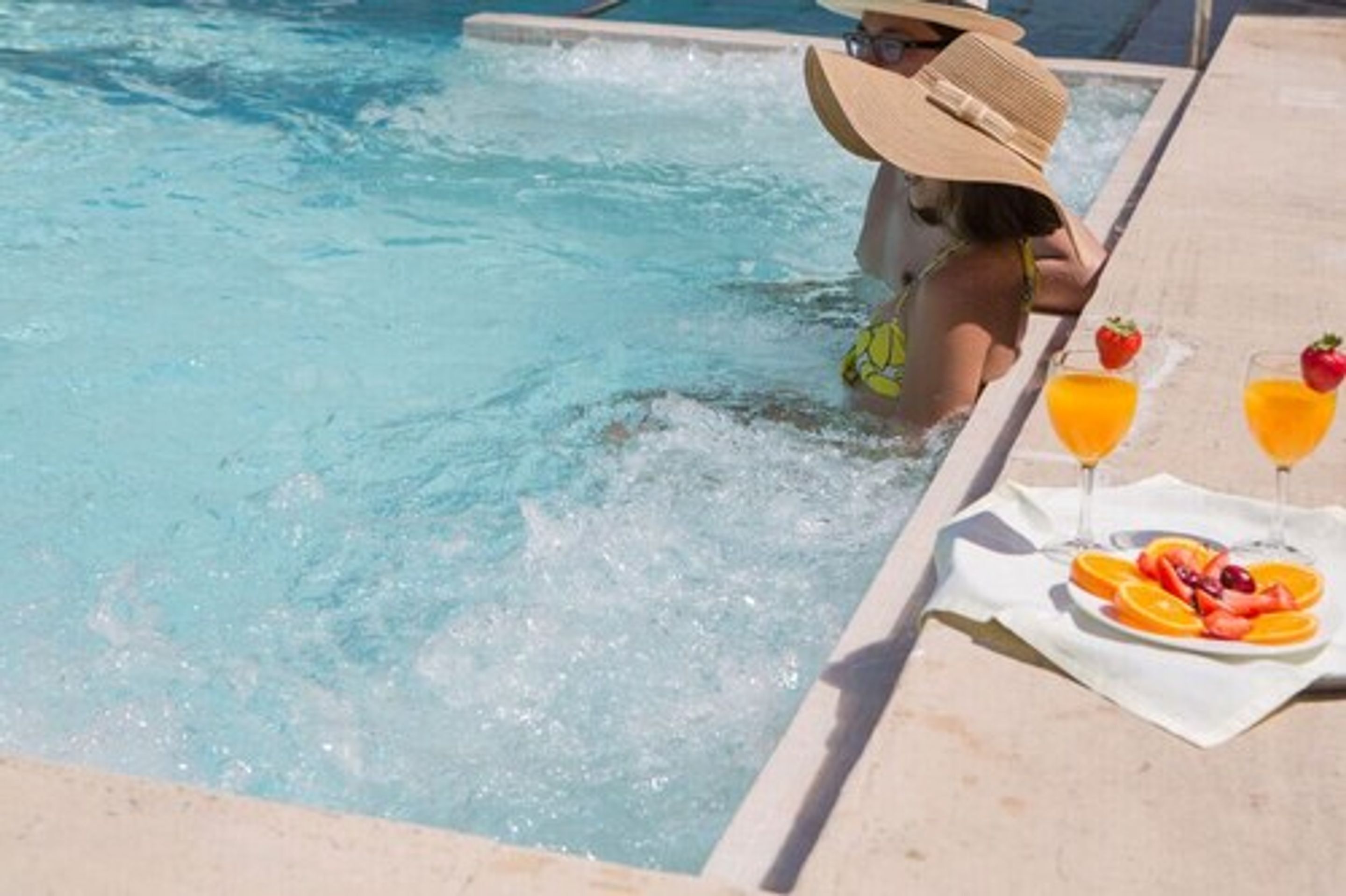 Area piscina con idromassaggio, tavolo con bevande e frutta, e una persona in costume da bagno e cappello.