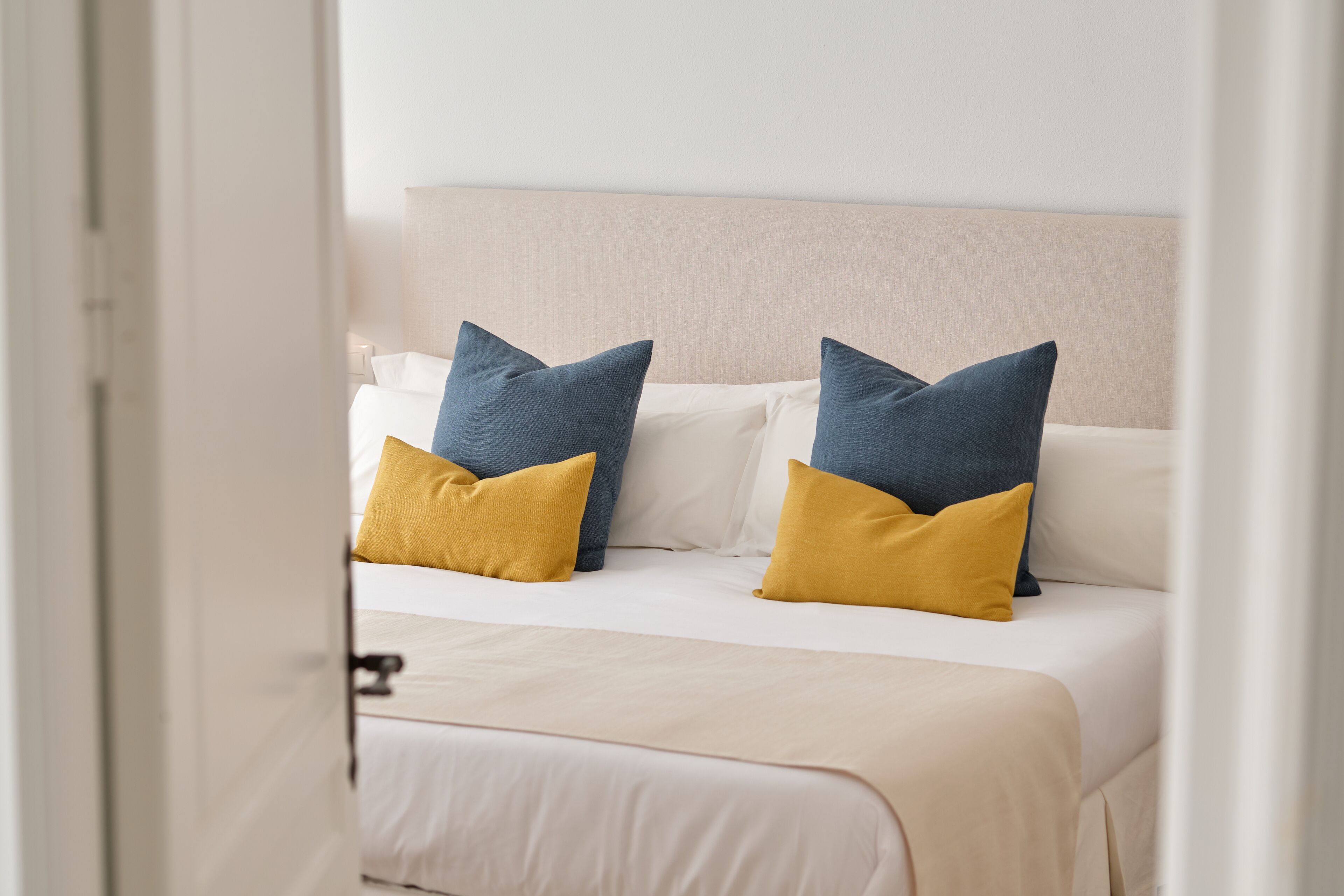 A neatly made bed with white and mustard bedding, two blue pillows, and a beige headboard.
