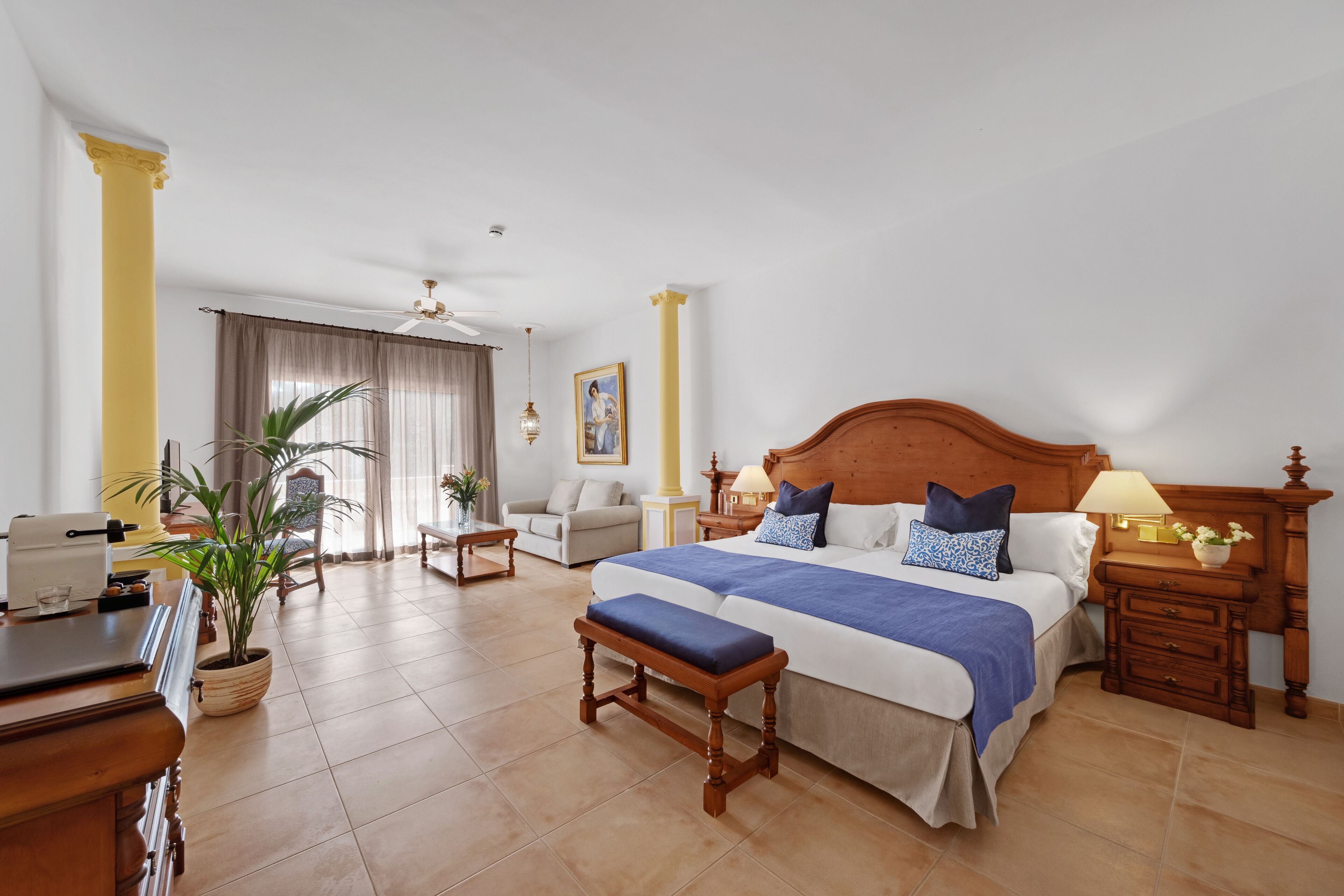 A hotel room with a large bed, a desk, a chair, a potted plant, and a window with curtains.