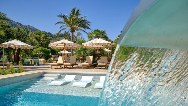 Un área de piscina con sillones de descanso y sombrillas, rodeada de palmeras y montañas.