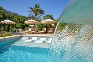 Un área de piscina con sillones de descanso y sombrillas, rodeada de palmeras y montañas.