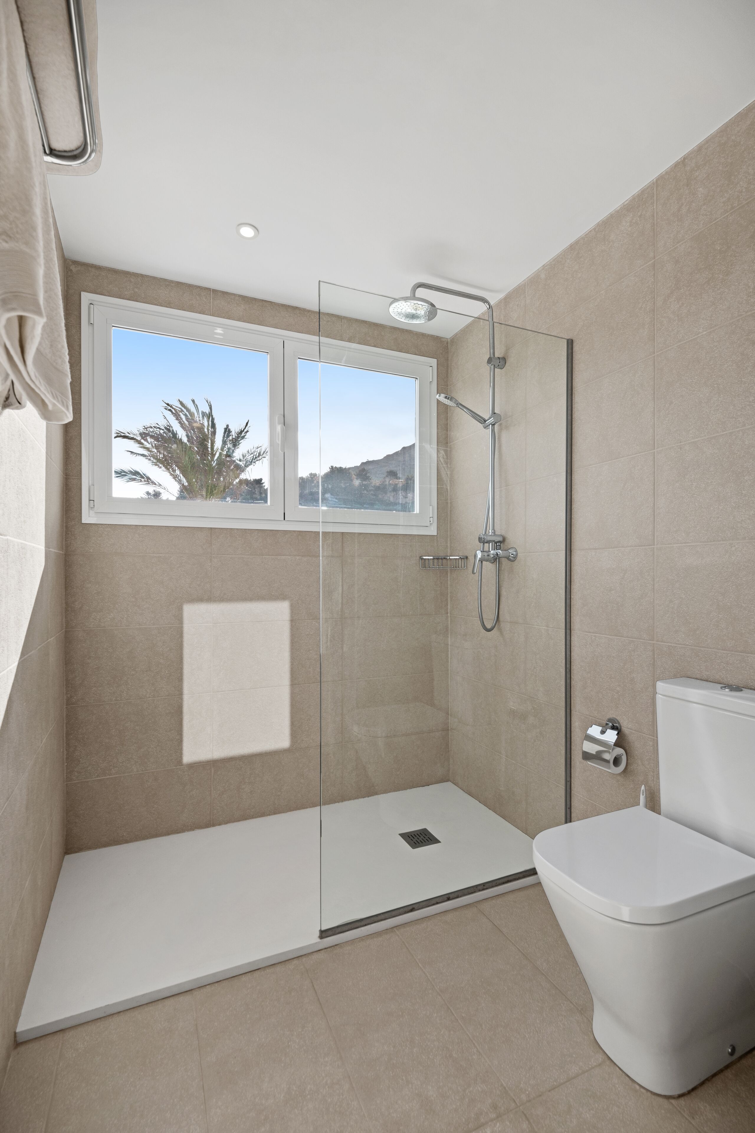 Un baño moderno con ducha de vidrio, inodoro y ventana con vista a palmeras y colinas.
