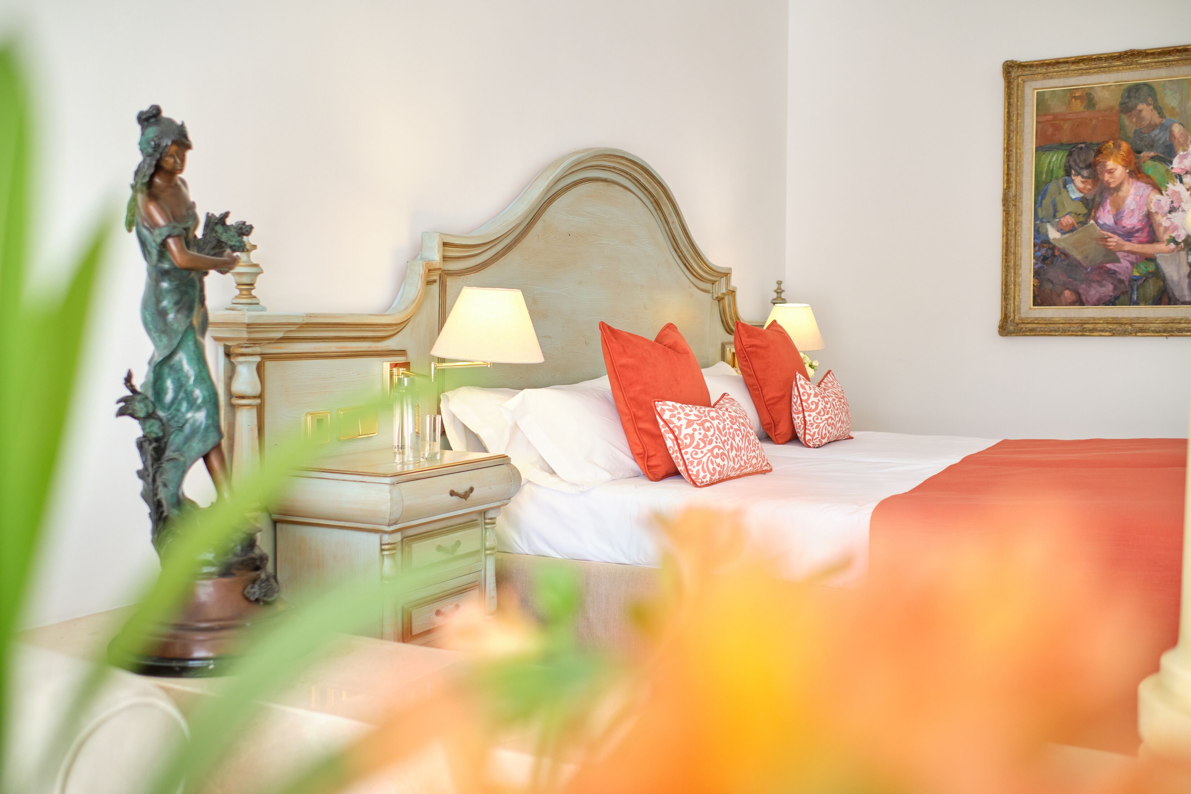 Un dormitorio con cama, almohadas, mesita de noche, lámpara y una estatua.